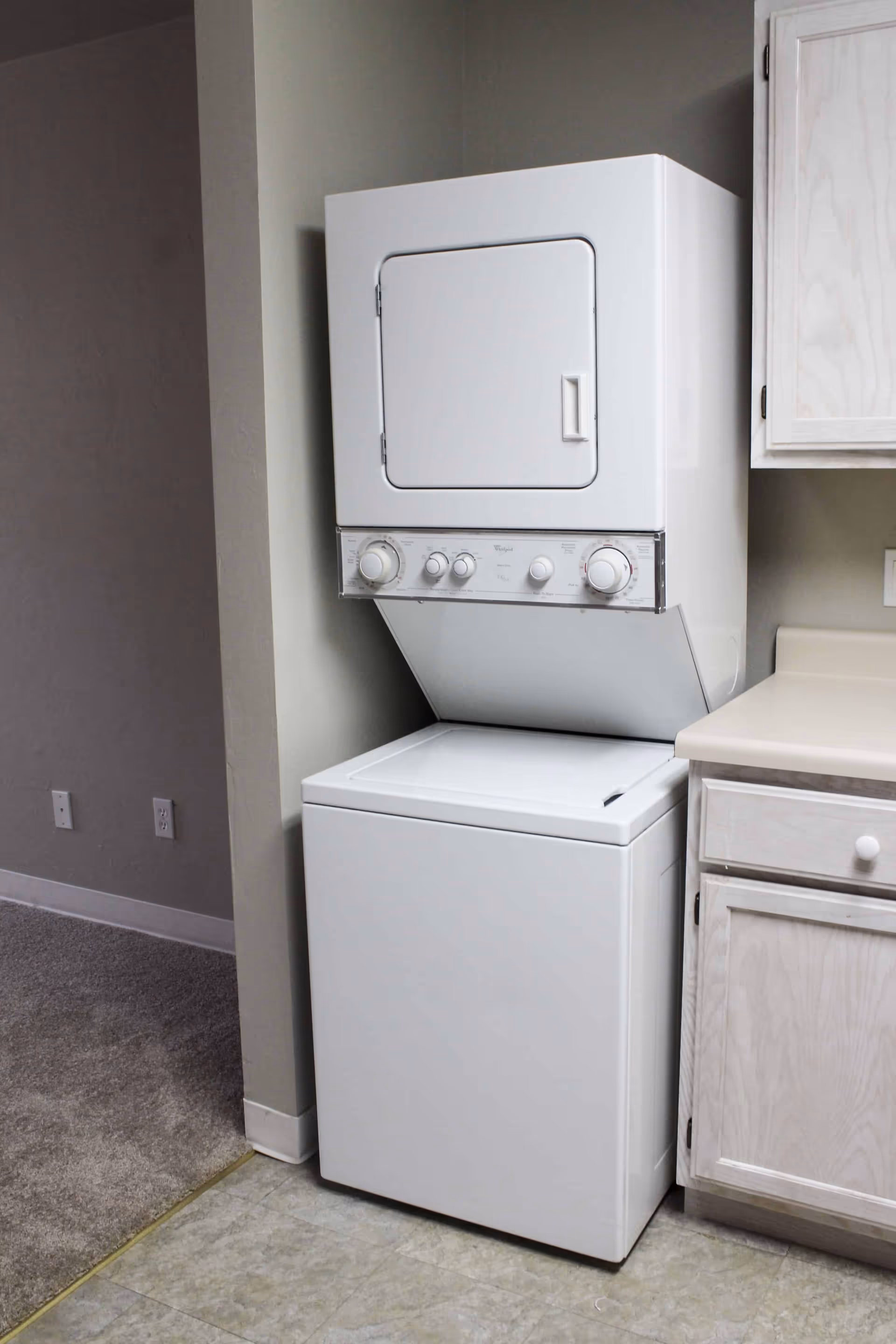 A white stacked washer and dryer unit placed in a corner next to a kitchen counter with light-colored cabinets. The floor is tiled and there is carpet visible in the adjacent room.