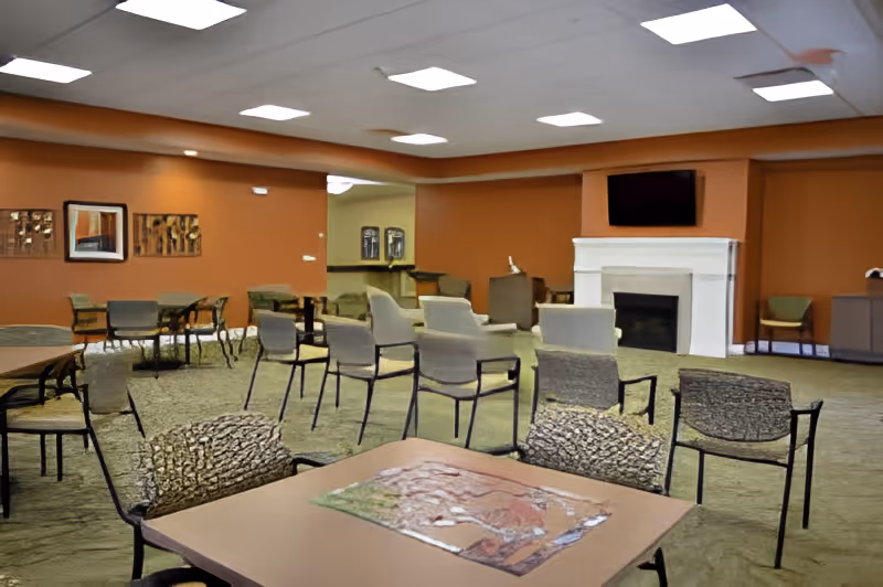 A spacious common area in a senior living facility with multiple tables and chairs arranged throughout the room. The walls are painted a warm orange color, and there is a white fireplace with a flat-screen TV mounted above it. The ceiling has recessed lighting, and there are framed artworks on the walls. The room has a carpeted floor and appears well-lit and inviting.