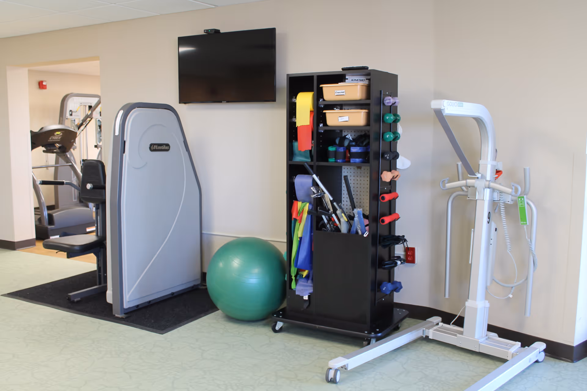 A small fitness area in a senior living facility featuring exercise equipment including a seated workout machine, a green exercise ball, a black storage rack with various exercise bands and weights, and a white patient lift device. A flat-screen TV is mounted on the wall above the equipment.