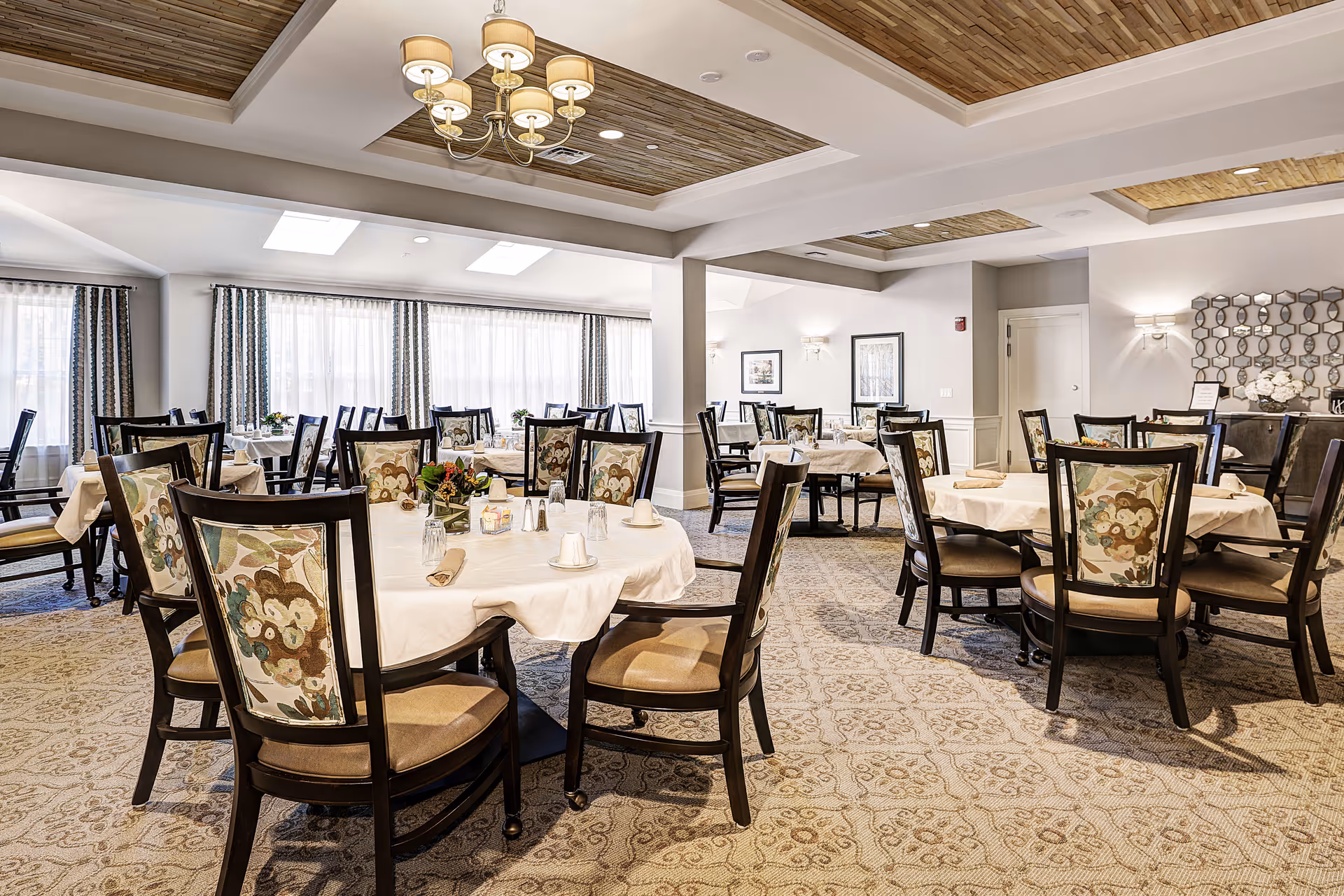 A bright and elegant dining room with multiple round tables covered with white tablecloths. Each table is surrounded by dark wooden chairs with floral patterned backs and beige cushions. The room features large windows with sheer curtains, a patterned carpet, and a ceiling with recessed wooden panels and a chandelier.