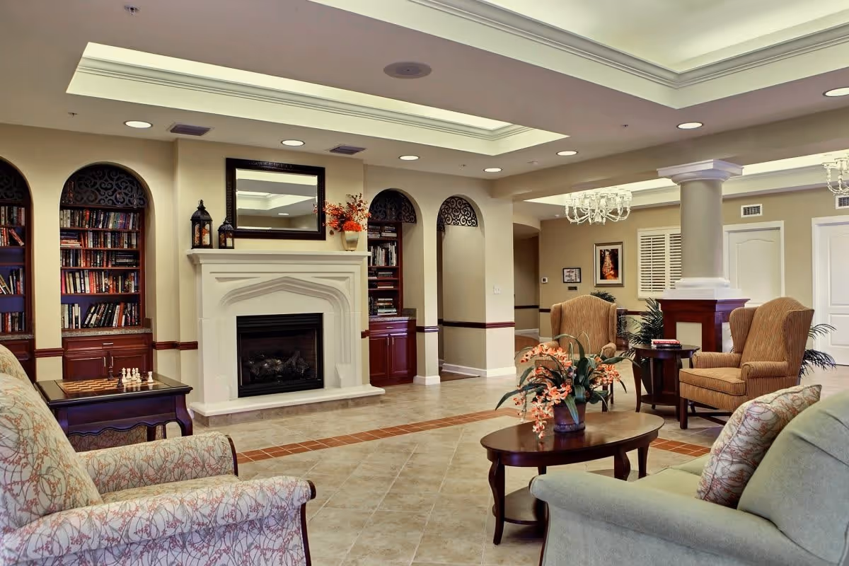 A spacious and elegant living room in an assisted living community featuring a white fireplace with a mirror above it, built-in bookshelves filled with books, comfortable upholstered armchairs and sofas, a wooden coffee table with a floral arrangement, and decorative columns under a coffered ceiling with recessed lighting.