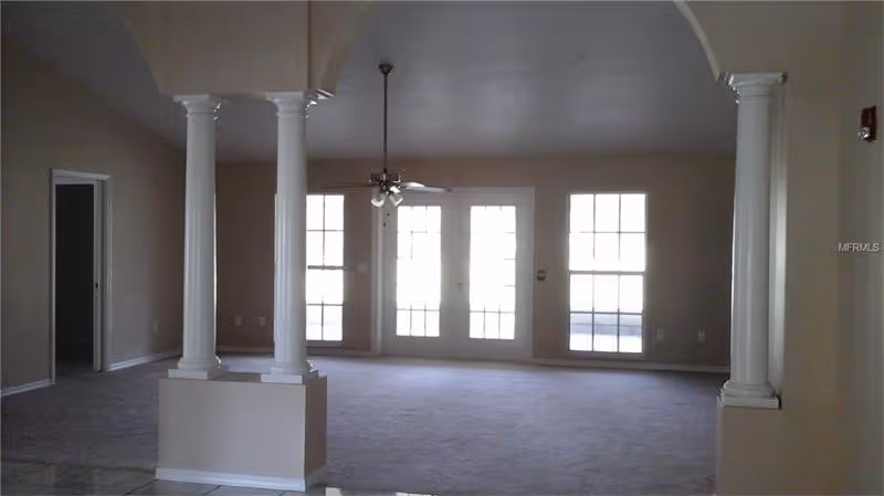 Spacious empty living room with carpet, decorative columns, a ceiling fan, and bright French doors and windows.