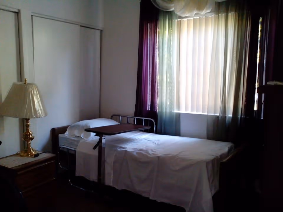 A simple bedroom with a single bed covered in white sheets and a pillow, a bedside table with a brass lamp, a window with vertical blinds and sheer curtains, and a wooden piece of furniture partially visible on the right.