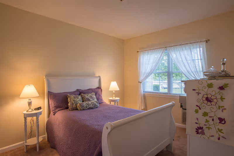 A cozy bedroom with a white wooden bed frame and a purple quilted bedspread. There are decorative pillows on the bed. Two white nightstands with lamps flank the bed. A window with sheer white curtains lets in natural light. A dresser with a floral embroidered cloth and glass decorative items is visible on the right side.