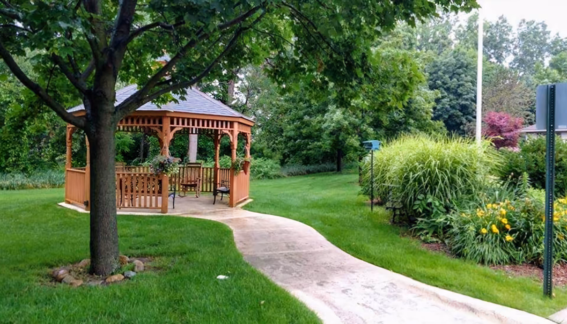 A wooden gazebo with a shingled roof situated in a green garden area. A concrete pathway curves toward the gazebo, which has hanging flower baskets and benches inside. Surrounding the gazebo are trees, bushes, and well-maintained grass.