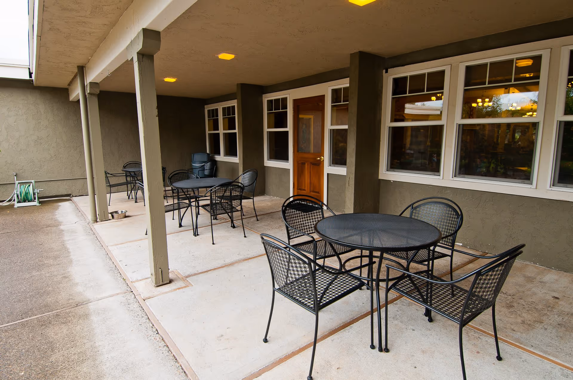 Covered outdoor patio area with several black metal tables and chairs arranged for seating. The patio is adjacent to a building with multiple windows and a wooden door. There is a garden hose reel on the left side and some stacked chairs in the back corner.