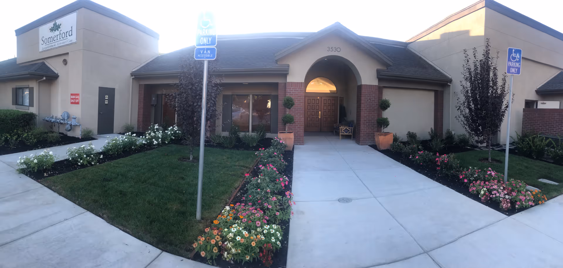 Exterior view of a single-story building with a beige facade and a dark roof. The entrance features a double wooden door under an arched portico with the number 3530 above it. There are two potted plants flanking the entrance and flower beds with colorful flowers and small trees on either side of the concrete walkway leading to the door. Two blue handicap parking signs are visible near the walkway. A sign on the left side of the building reads 'Somerford Assisted Living & Alzheimer's Care.'