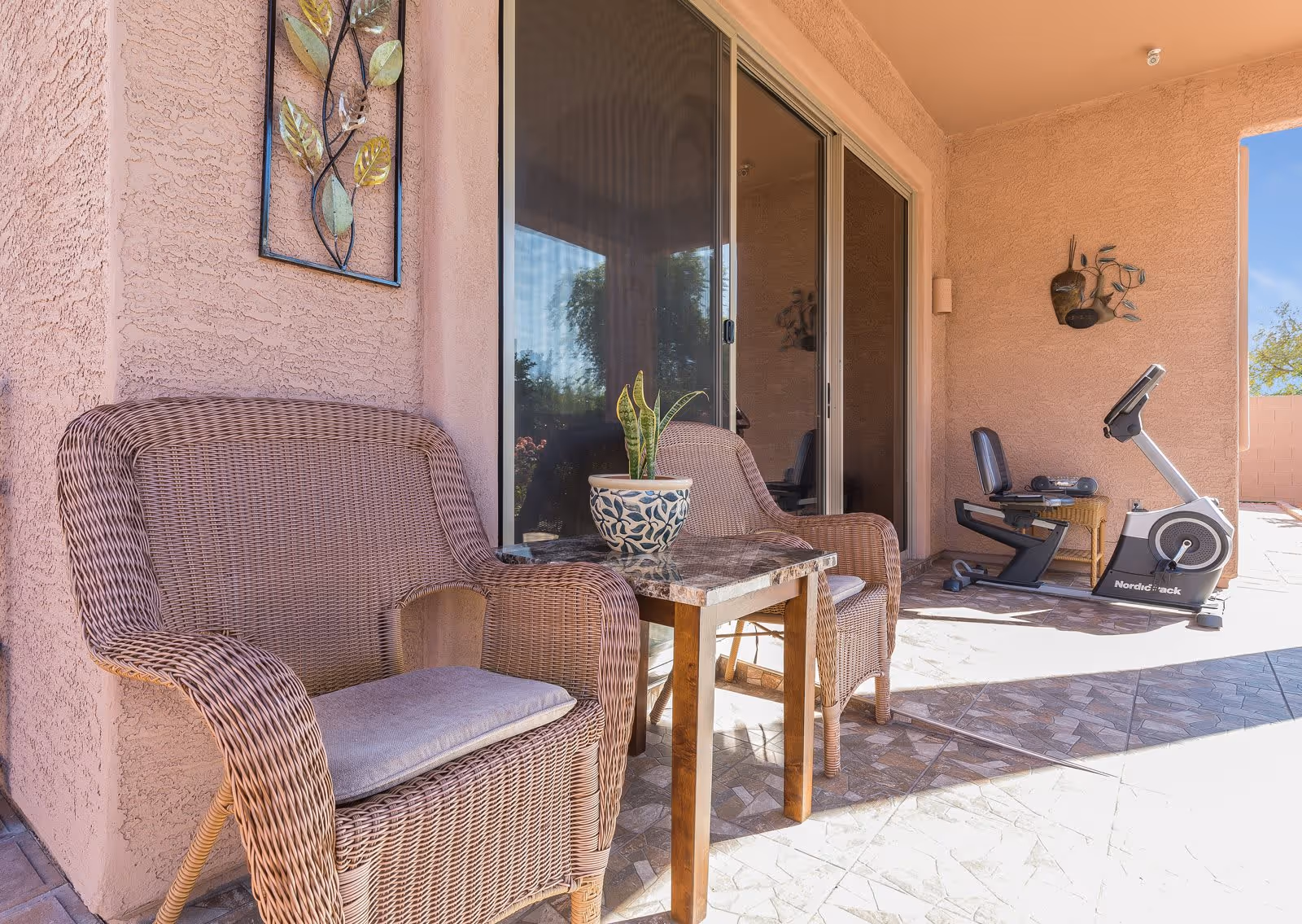 Outdoor patio area with two wicker chairs and a small table with a potted plant. There is a stationary exercise bike and a small wicker side table with a radio on it. The patio has tiled flooring and peach-colored stucco walls with decorative metal wall art. Sliding glass doors lead inside the building.
