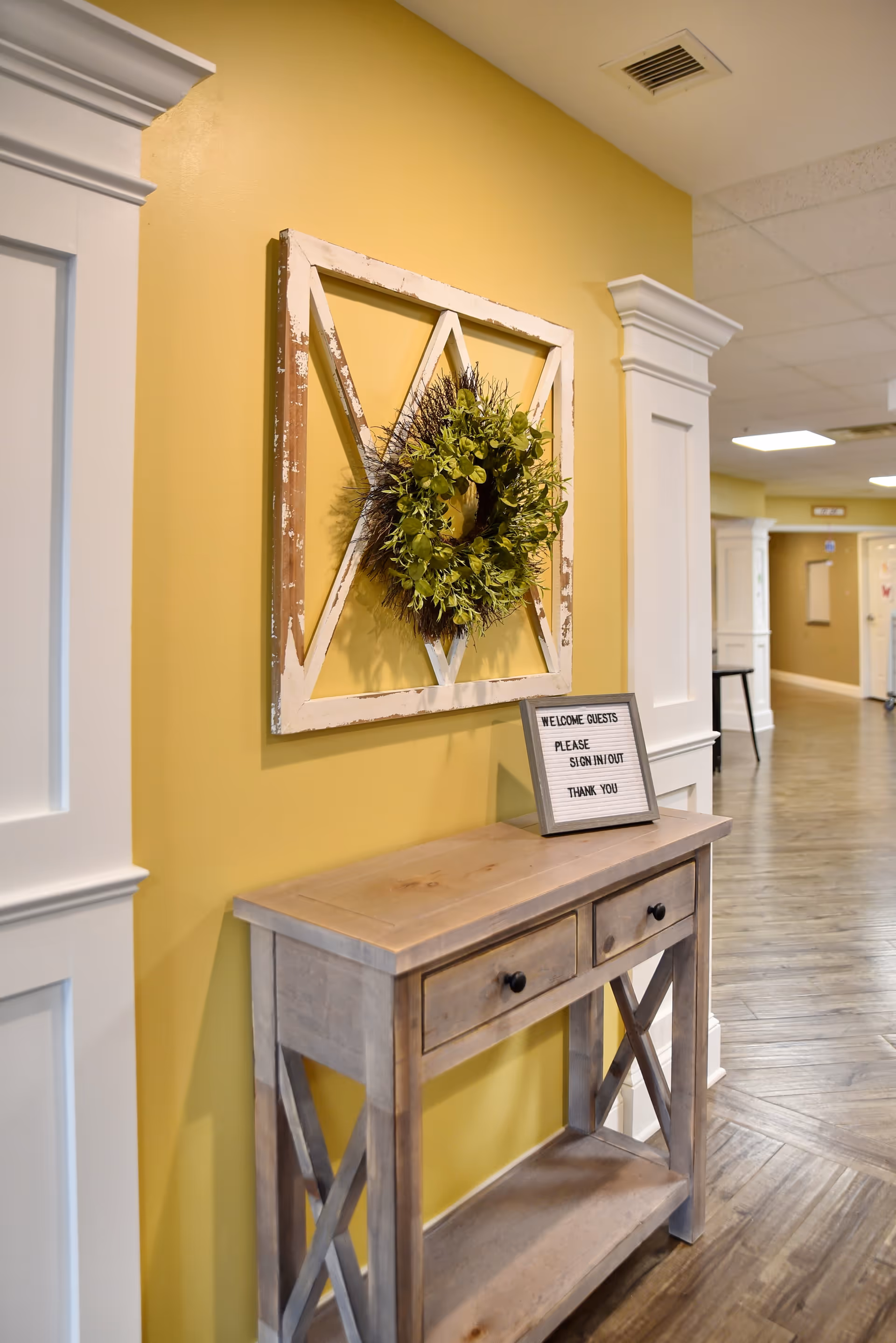 A hallway corner in a senior living facility with a wooden console table against a yellow wall. Above the table is a decorative wooden frame with a green wreath. On the table is a small sign that reads 'WELCOME GUESTS PLEASE SIGN IN/OUT THANK YOU'. The hallway has wood-patterned flooring and white trim around the doors and columns.