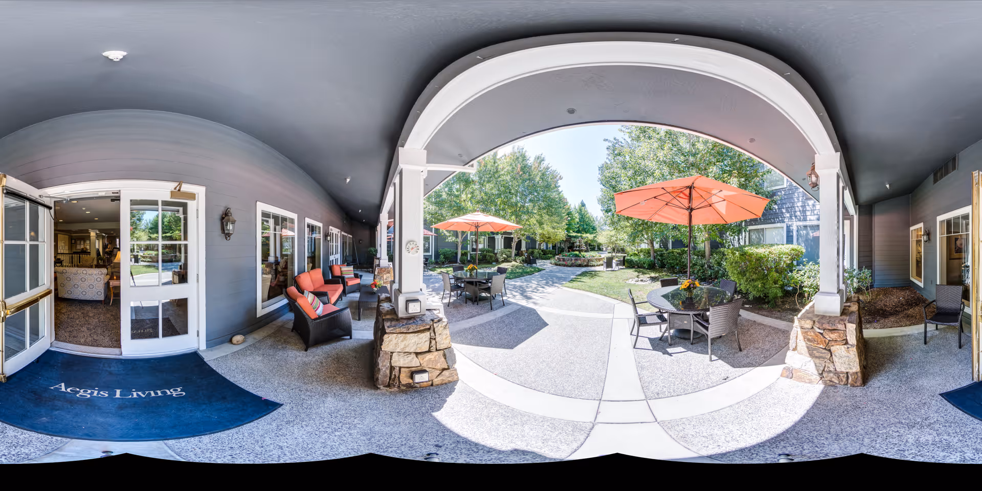 Covered outdoor patio area with seating including cushioned chairs and tables with orange umbrellas, leading to a garden with trees and shrubs. The entrance to the building is visible with a blue mat that reads 'Aegis Living'.