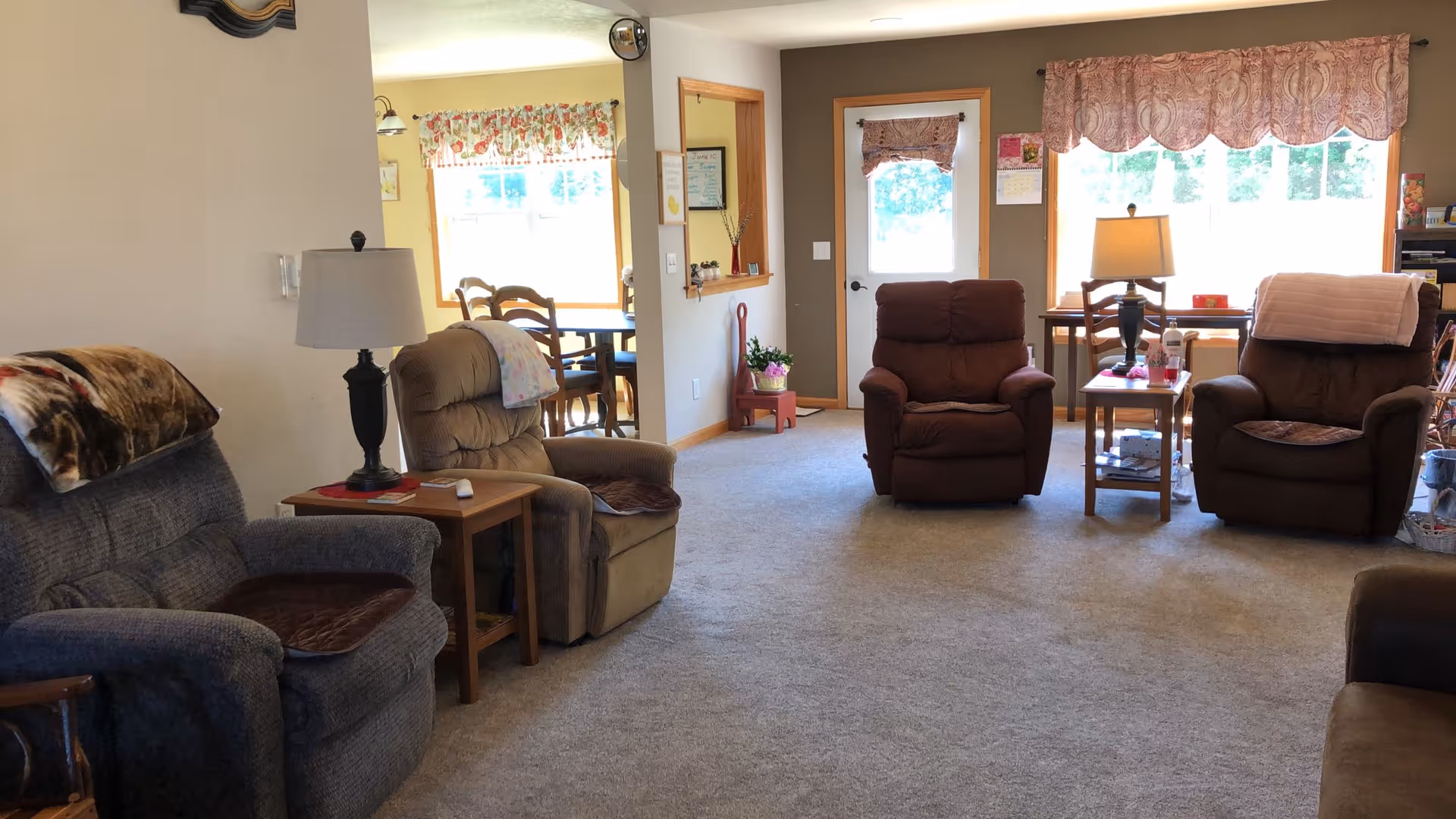 Communal living room with several recliners, side tables and lamps facing large windows and a door.