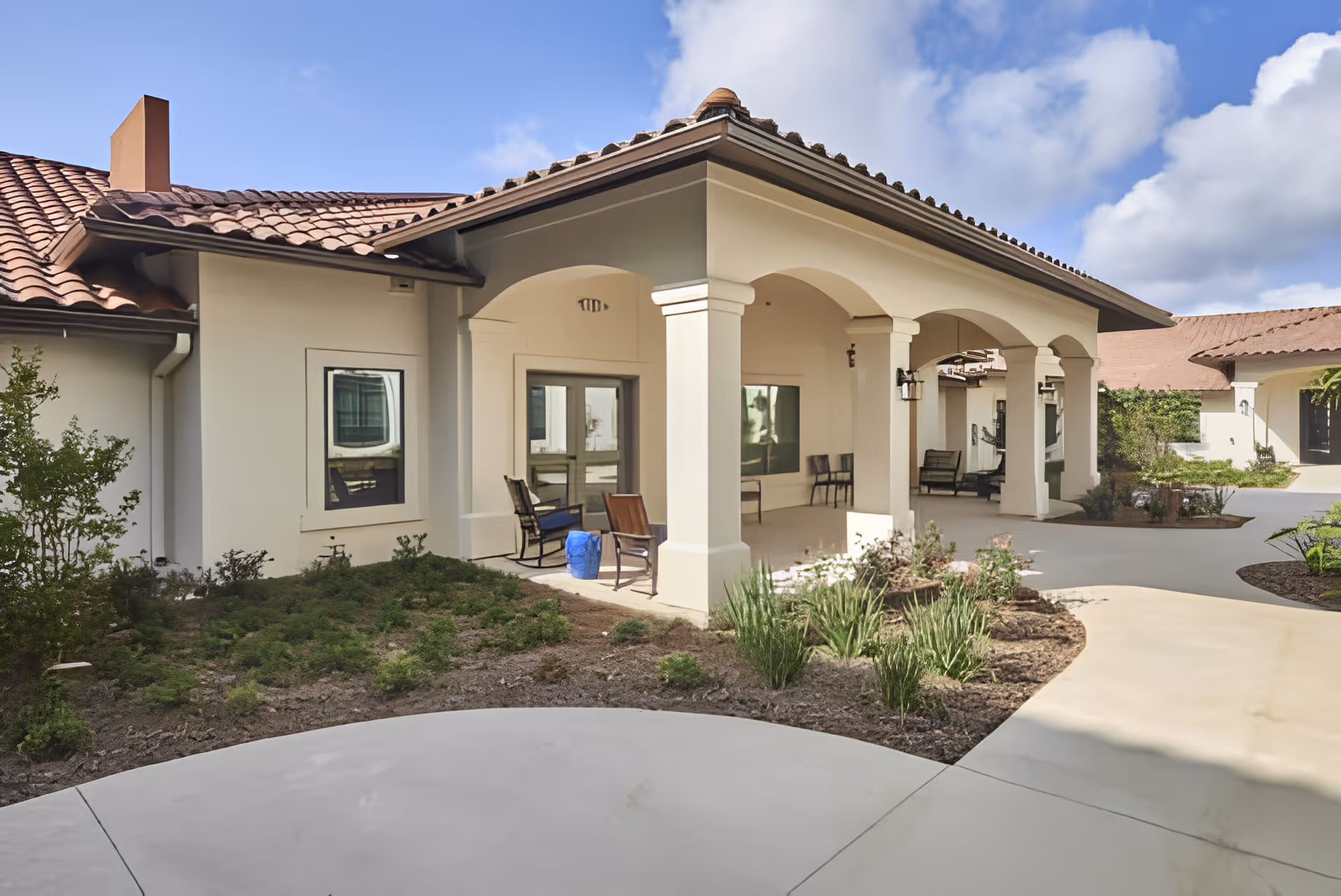 Outdoor view of a senior living facility with a covered patio area featuring several chairs. The building has a tiled roof and light-colored walls, with landscaping including bushes and plants around the paved walkway.