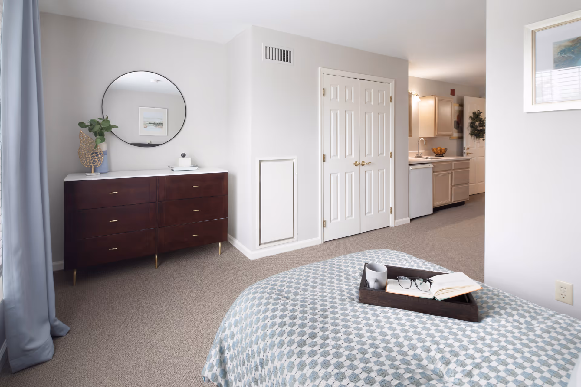A bright and clean bedroom area with a patterned bedspread, a wooden tray holding a mug, glasses, and an open book on the bed. There is a dark wooden dresser with a round mirror above it, a small plant, and decorative items. In the background, a kitchen area with light-colored cabinets and a dishwasher is visible. The walls are painted light gray, and the floor is carpeted.