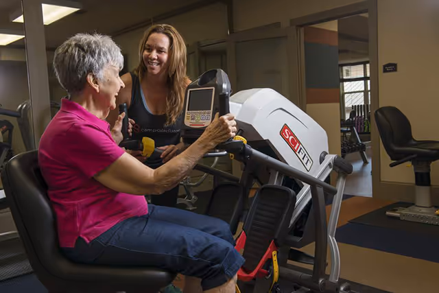 An elderly woman in a pink shirt and blue pants is using a seated exercise machine in a fitness room, while a younger woman in athletic wear smiles and encourages her.