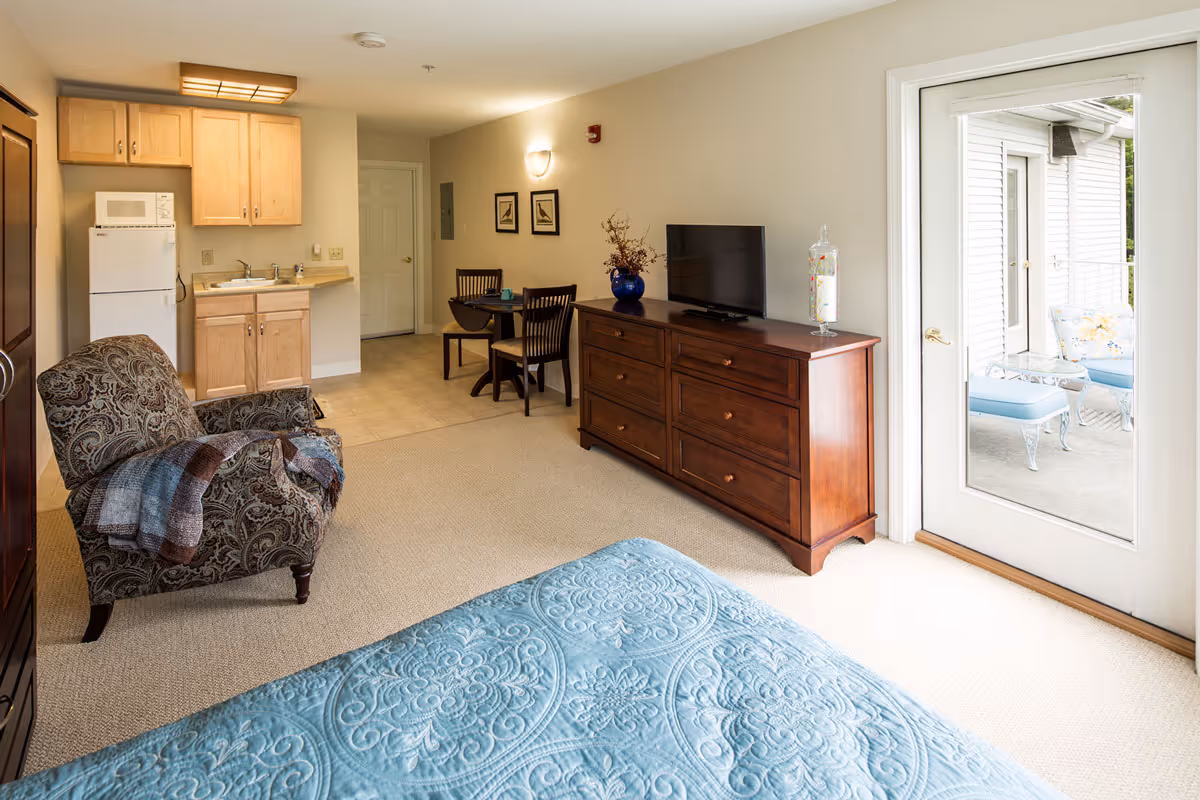 A cozy assisted living room featuring a patterned armchair with a blanket, a wooden dresser with a flat-screen TV and decorative items, a small dining table with two chairs, a kitchenette with a refrigerator, microwave, and sink, and a bed with a blue quilt. A glass door leads to an outdoor patio with white metal furniture and blue cushions.
