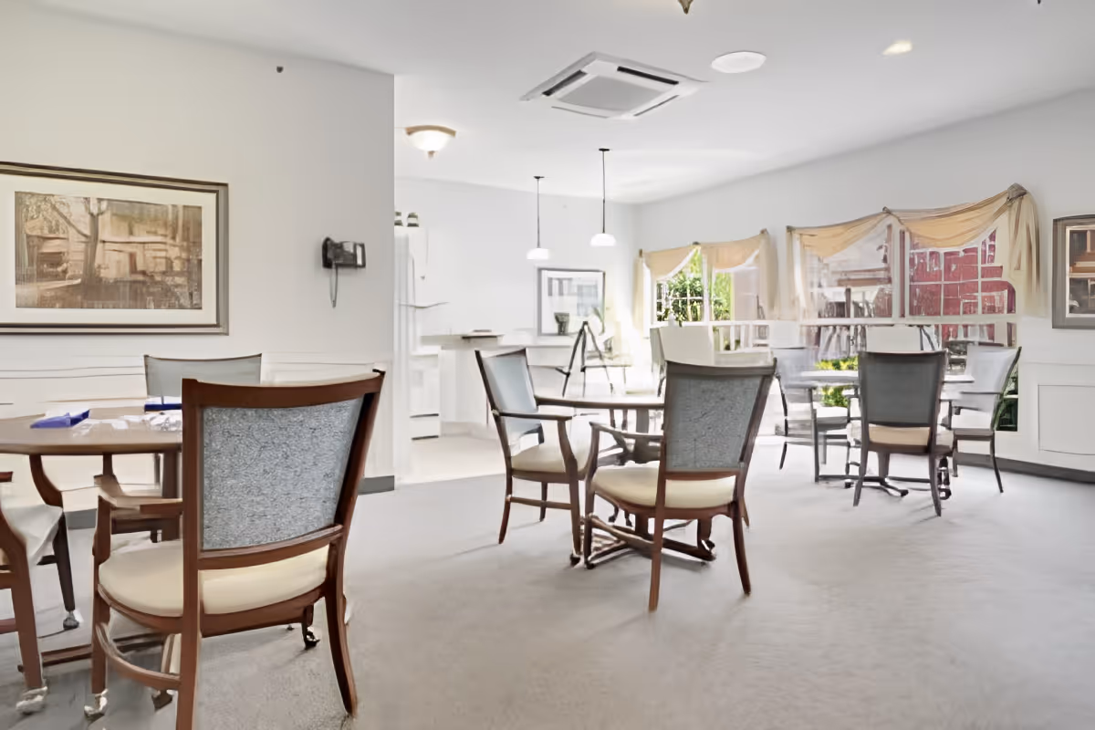 A bright dining area in a senior living facility with several round wooden tables and cushioned chairs. Large windows with sheer curtains allow natural light to fill the room. The walls are decorated with framed artwork, and pendant lights hang from the ceiling. The space appears clean and welcoming.