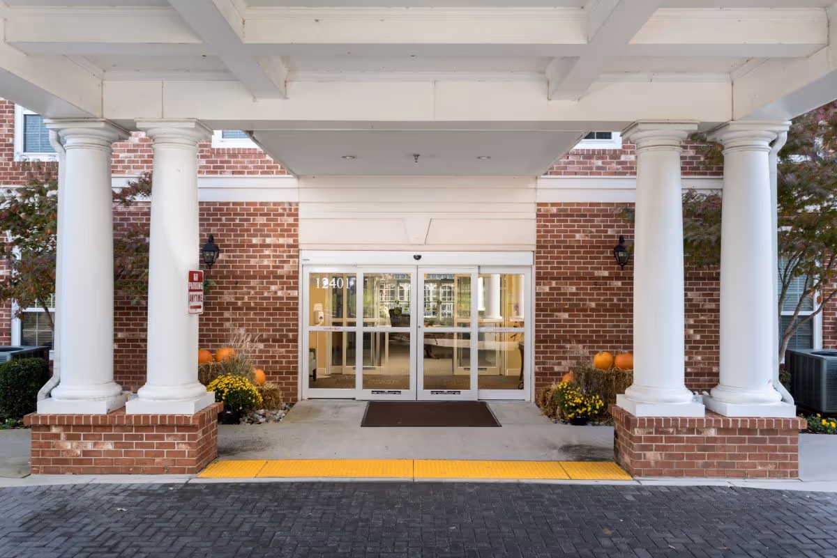 Entrance to a building with large white columns on brick bases, automatic glass sliding doors, and a covered driveway. There are pumpkins and yellow flowers on either side of the entrance, and a yellow tactile paving strip on the ground in front of the door.