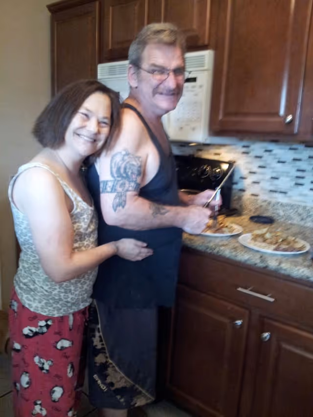 A man and a woman standing together in a kitchen. The man is cutting food on a plate placed on the kitchen counter, and the woman is standing behind him, smiling and hugging him around the waist. The kitchen has wooden cabinets, a microwave, and a tiled backsplash.