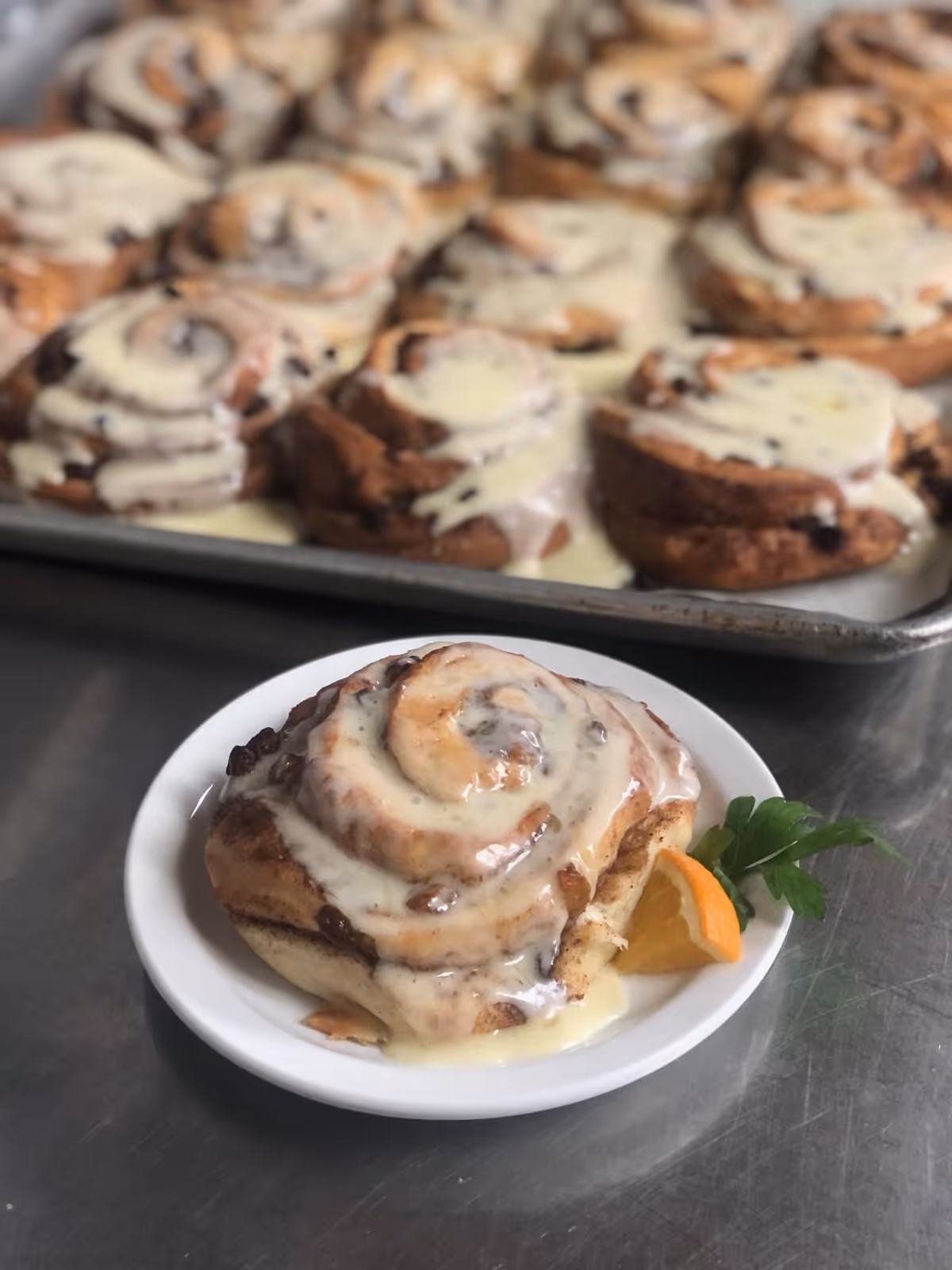 A close-up of a cinnamon roll with icing on a white plate, garnished with a small orange slice and a sprig of parsley. In the background, there is a tray filled with more cinnamon rolls covered in icing.