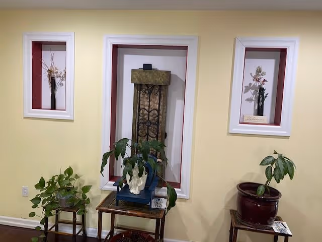 Yellow interior wall with three recessed framed niches holding decorative floral arrangements and a central niche with a decorative panel above small tables and potted plants.