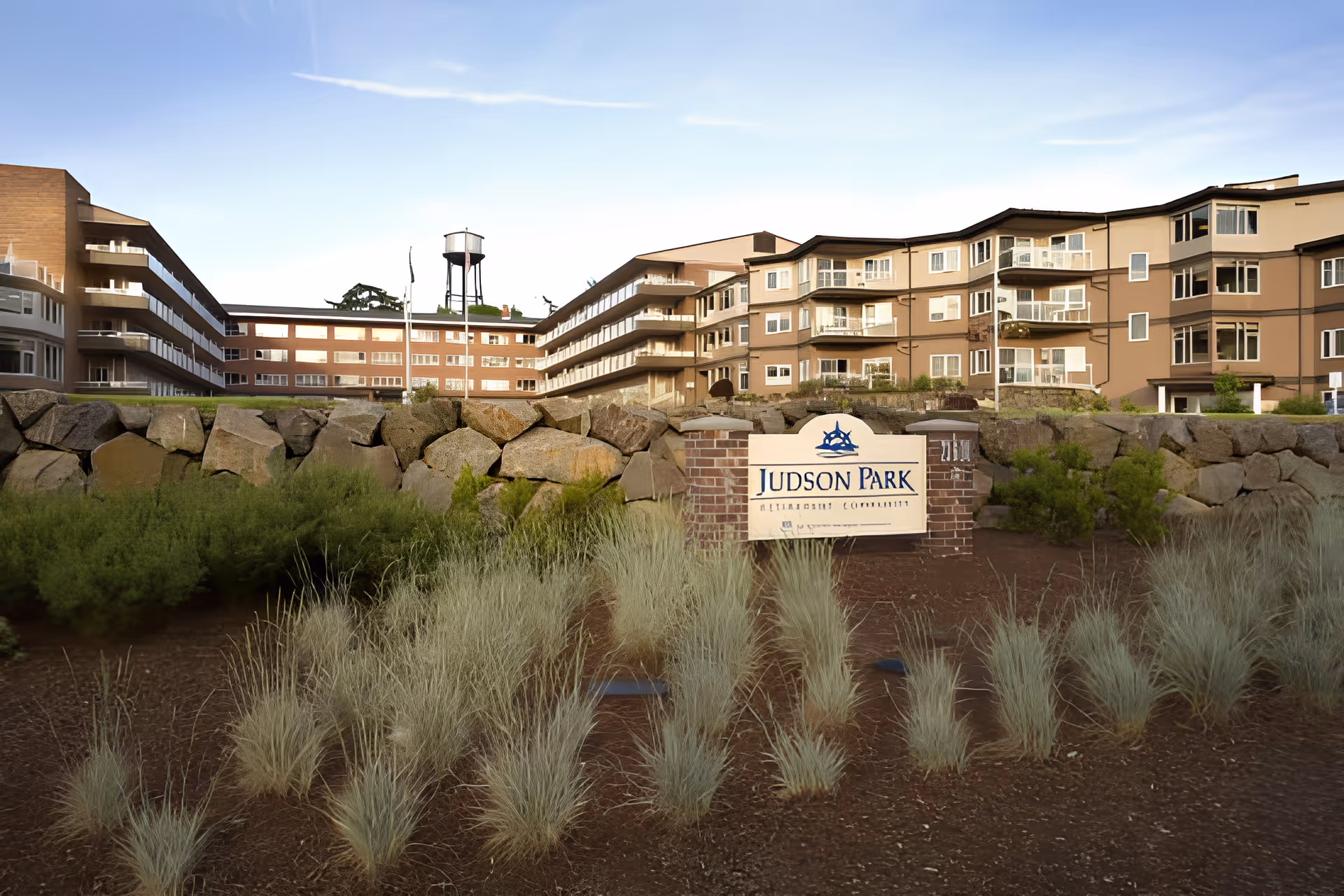 Exterior view of Judson Park Retirement Community building with multiple floors and balconies, a stone wall, and landscaped plants in the foreground under a clear sky.