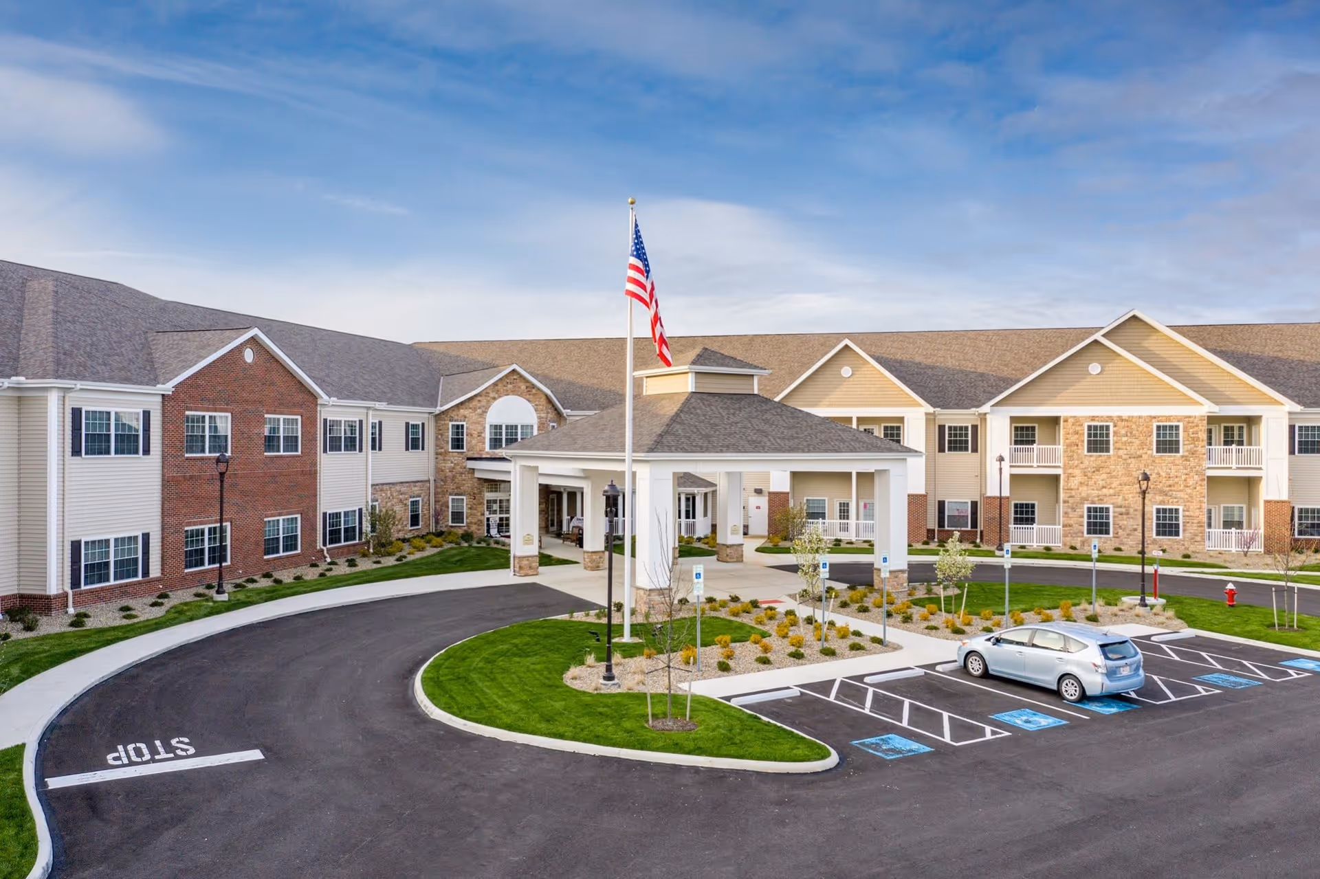 Front exterior view of a senior living facility named Danbury North Ridgeville with a circular driveway, an American flag on a flagpole, landscaped greenery, and a parked car in a handicapped parking space.