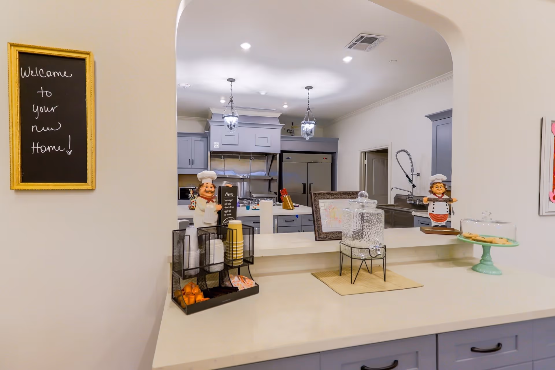 Service counter and pass-through to a clean commercial-style kitchen with decorative chef figurines, a beverage dispenser, and a 'Welcome to your new Home!' chalkboard.