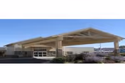 Exterior view of a single-story building with a large covered entrance and a clear blue sky. The building has beige walls and stone accents near the entrance, with some landscaping including bushes and plants in front.