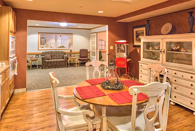 A cozy dining area with a round wooden table set with four chairs and red placemats. A decorative lantern centerpiece is on the table. To the right, there is a white wooden cabinet with glass doors displaying decorative items. In the background, an adjoining living room area is visible with a patterned sofa, a chair, side tables, and a lamp. The walls are painted in warm tones, and the floor is a combination of wood and carpet.