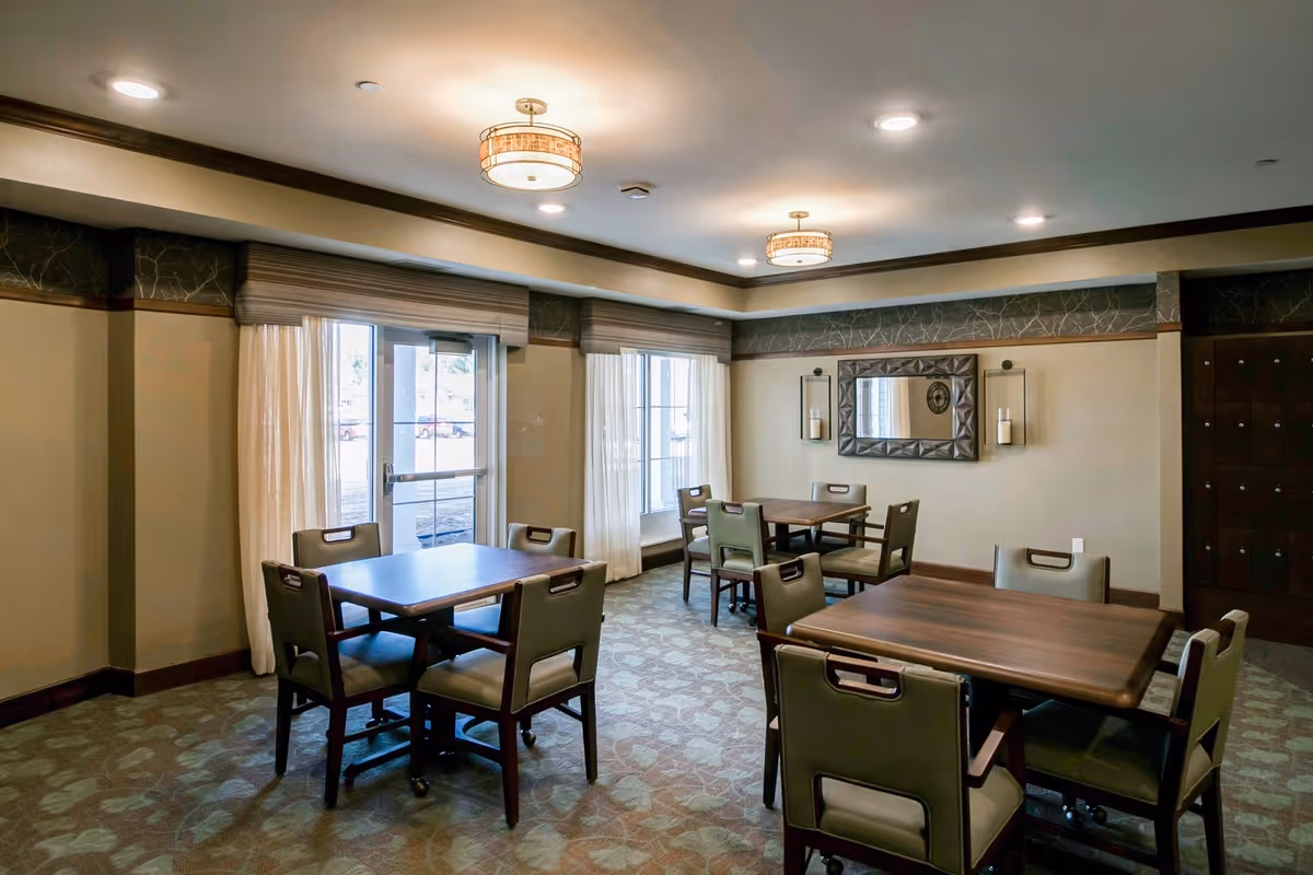 A well-lit room with three wooden tables, each surrounded by four green cushioned chairs. The room has large windows with sheer white curtains and beige walls decorated with a large mirror and wall sconces. The ceiling features recessed lighting and two decorative light fixtures.