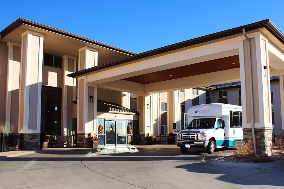 Front entrance of the Pointe Frontier senior living building with a covered porte-cochere and a white-and-teal shuttle van parked nearby.