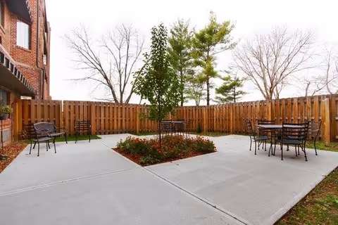 Outdoor patio area with concrete flooring, a small tree planted in the center surrounded by flowers, metal benches and tables with chairs, enclosed by a wooden fence. Bare trees and a brick building are visible in the background.
