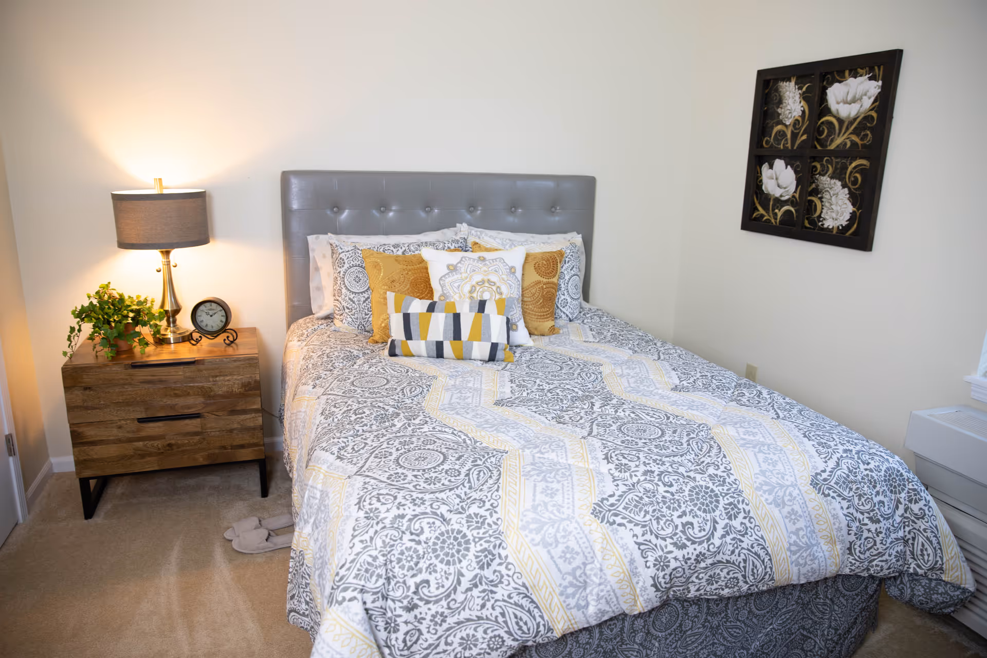 A neatly made bed with a gray tufted headboard, patterned bedding in gray, white, and yellow, and several decorative pillows. To the left of the bed is a wooden nightstand with a lamp, a small clock, and a green plant. On the wall above the bed is a framed artwork featuring four floral designs. The room has beige carpeting and light-colored walls.