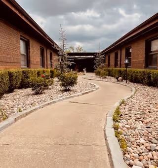 Curved concrete pathway between two single-story brick buildings with small bushes and rock landscaping on either side under a cloudy sky.
