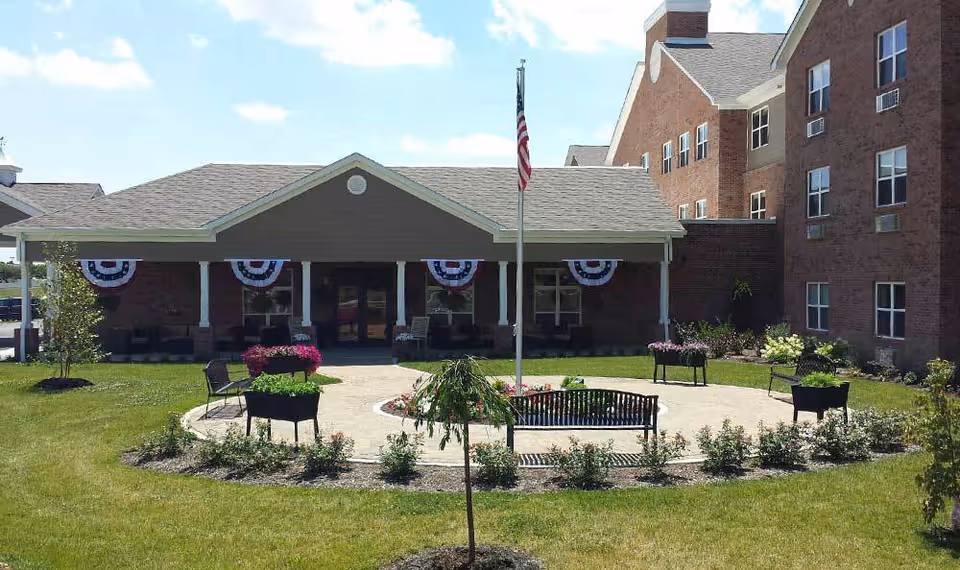 Front courtyard of a brick senior living building with a flagpole, benches, planters, and a covered entrance decorated with patriotic bunting.