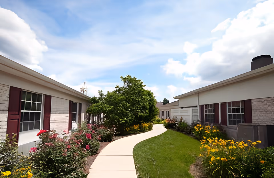 Curved paved walkway through a landscaped courtyard between single-story buildings with flower beds and a partly cloudy sky.