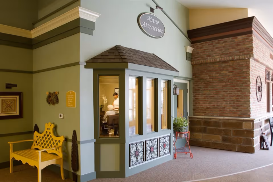 Interior hallway of a senior living facility featuring a small salon storefront labeled 'Mane Attraction', a yellow bench, and a decorative brick wall.