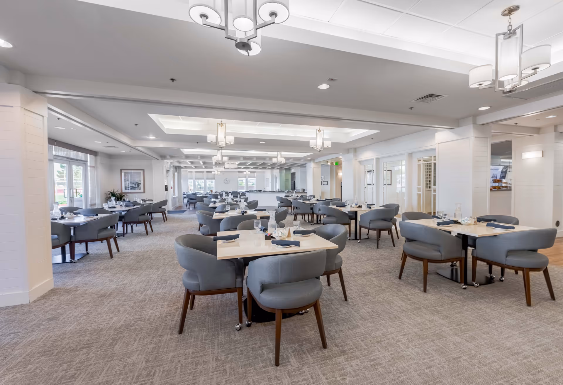 A spacious and well-lit dining room with multiple tables set with glasses, napkins, and cutlery. The room features modern gray chairs with wooden legs, light-colored walls, and large windows allowing natural light to enter. Ceiling lights and chandeliers provide additional illumination.