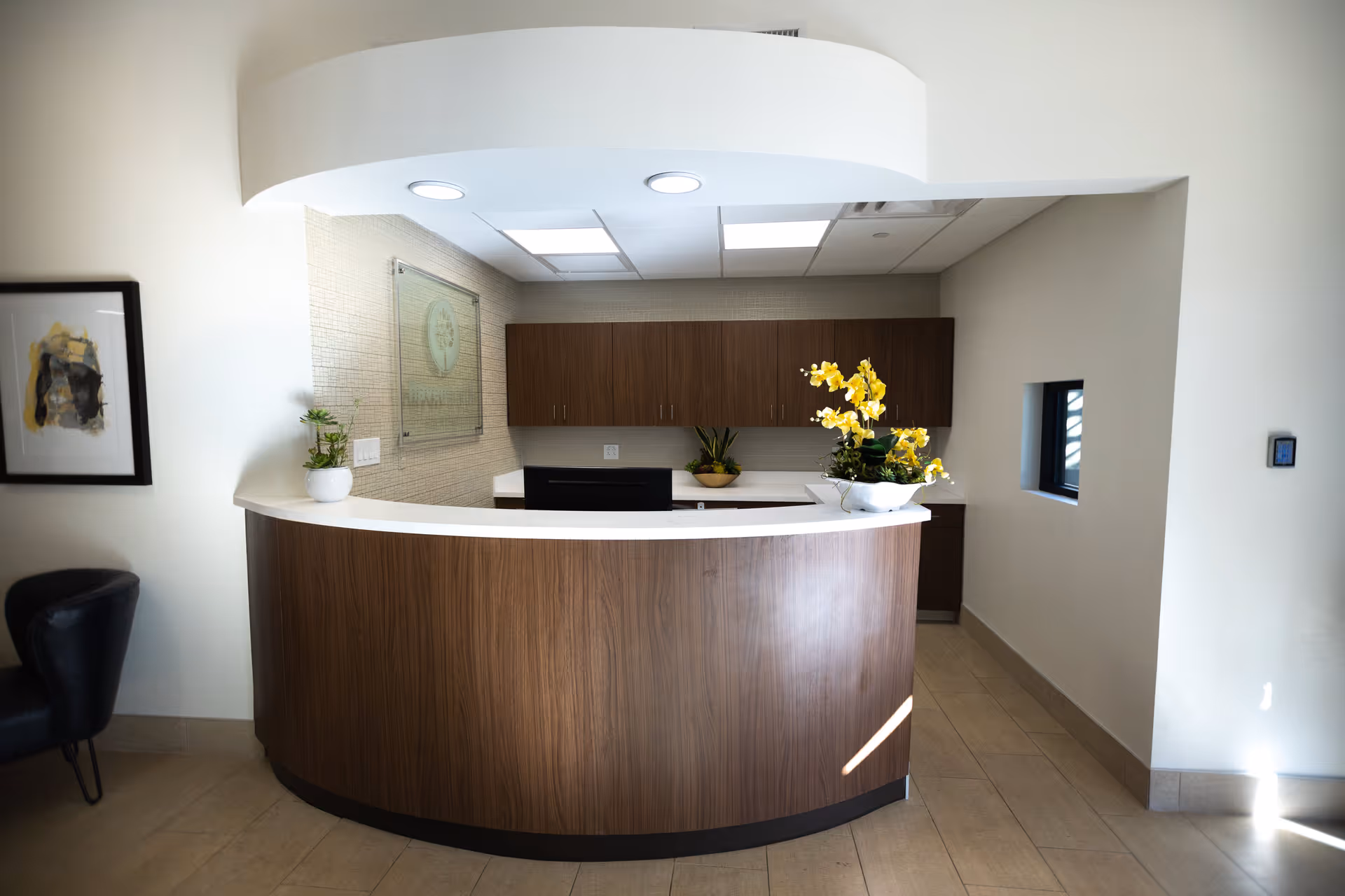 A modern reception desk area with a curved wooden front and white countertop. Behind the desk are wooden cabinets mounted on a beige tiled wall. The desk has a computer monitor and two flower arrangements with yellow flowers. To the left, there is a black chair and a framed abstract artwork on the wall. The floor is tiled and there is a small window on the right wall.