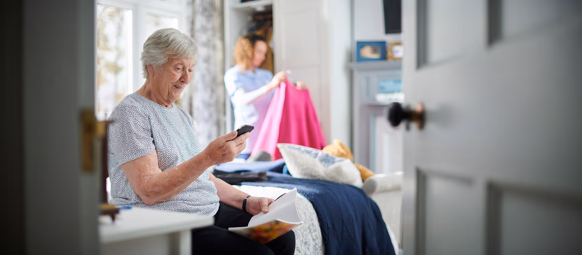 An older woman sits on a bed looking at her phone while another person in the background holds up a pink garment in a sunlit bedroom.