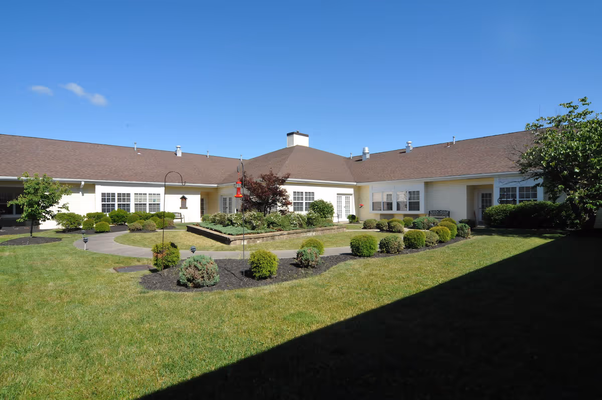 A sunny courtyard garden area at Celebration Villa of Chippewa featuring a well-maintained lawn, neatly trimmed bushes, a raised flower bed with various plants, and a bird feeder. The courtyard is surrounded by a single-story building with multiple windows and a brown roof under a clear blue sky.