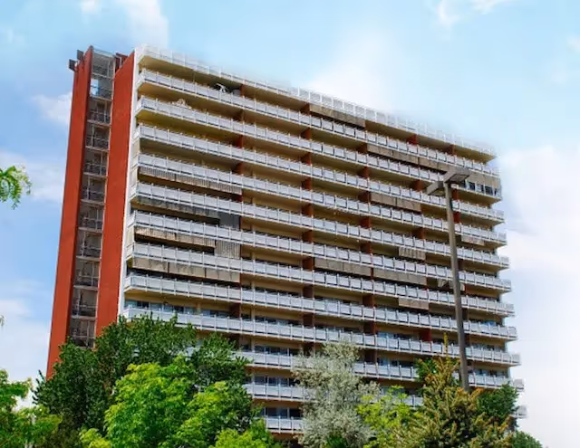 Tall multi-story assisted living center building with balconies on each floor, surrounded by green trees under a partly cloudy sky.