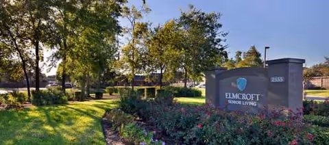 A landscaped entrance with a stone sign reading Elmcroft Senior Living beside trees.