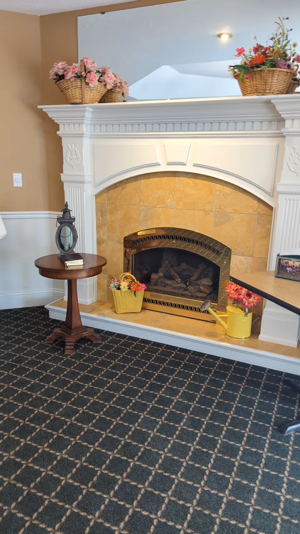 A cozy indoor fireplace with a white mantel and beige tile surround. On the mantel are two baskets filled with pink and orange flowers. In front of the fireplace is a small yellow basket with flowers and a yellow watering can with pink flowers. To the left of the fireplace is a small round wooden table with a decorative lantern and a small book on it. The floor is carpeted with a dark patterned carpet.