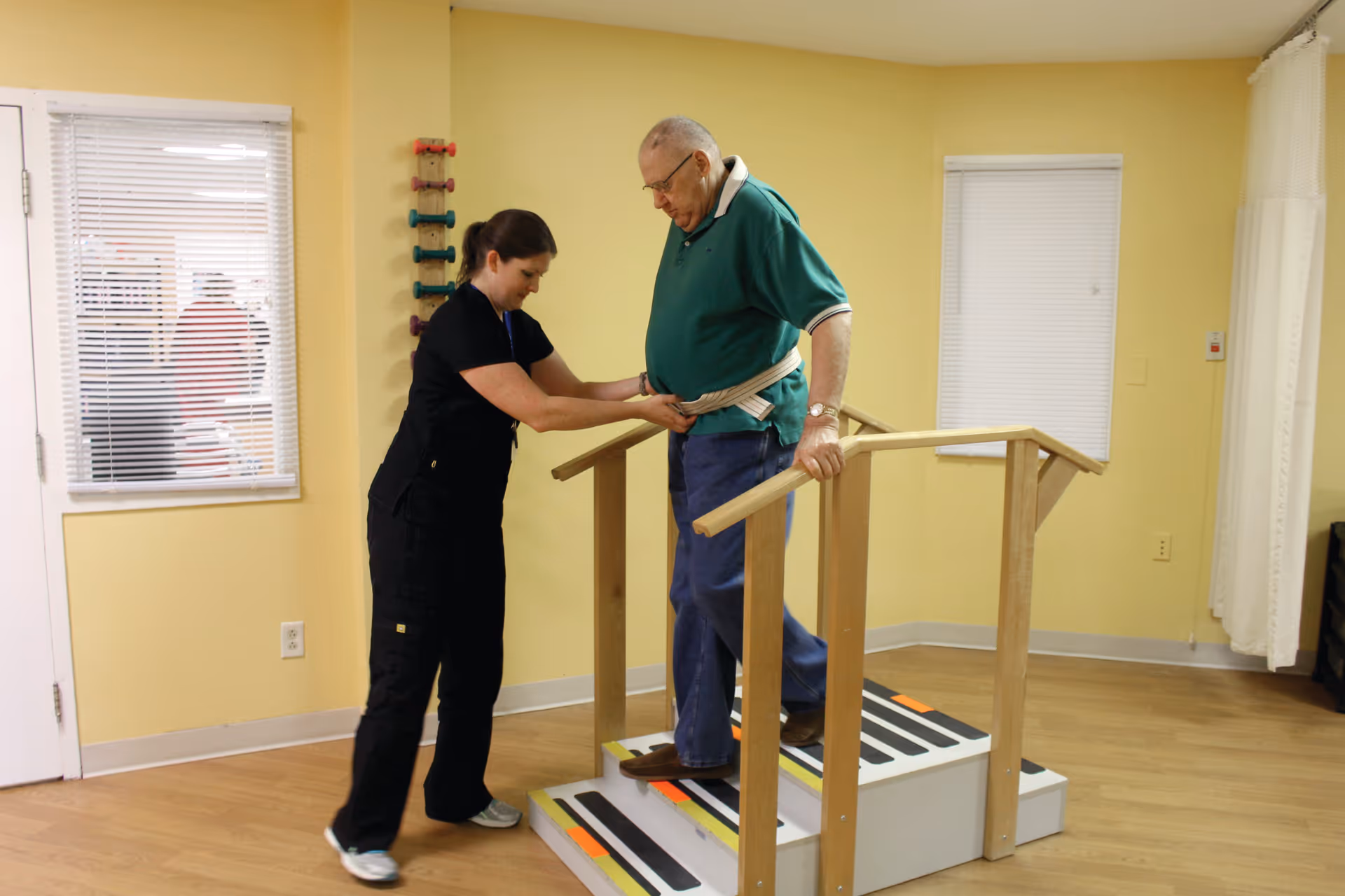 A healthcare worker assists an elderly man as he practices walking up and down a small set of stairs with handrails in a therapy or rehabilitation room with yellow walls and wooden flooring.
