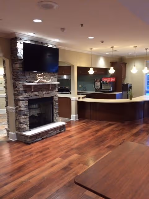 Interior view of a senior living facility common area featuring a stone fireplace with a mounted flat-screen TV above it, a curved wooden counter with pendant lights hanging overhead, a beverage dispenser, and wooden flooring.