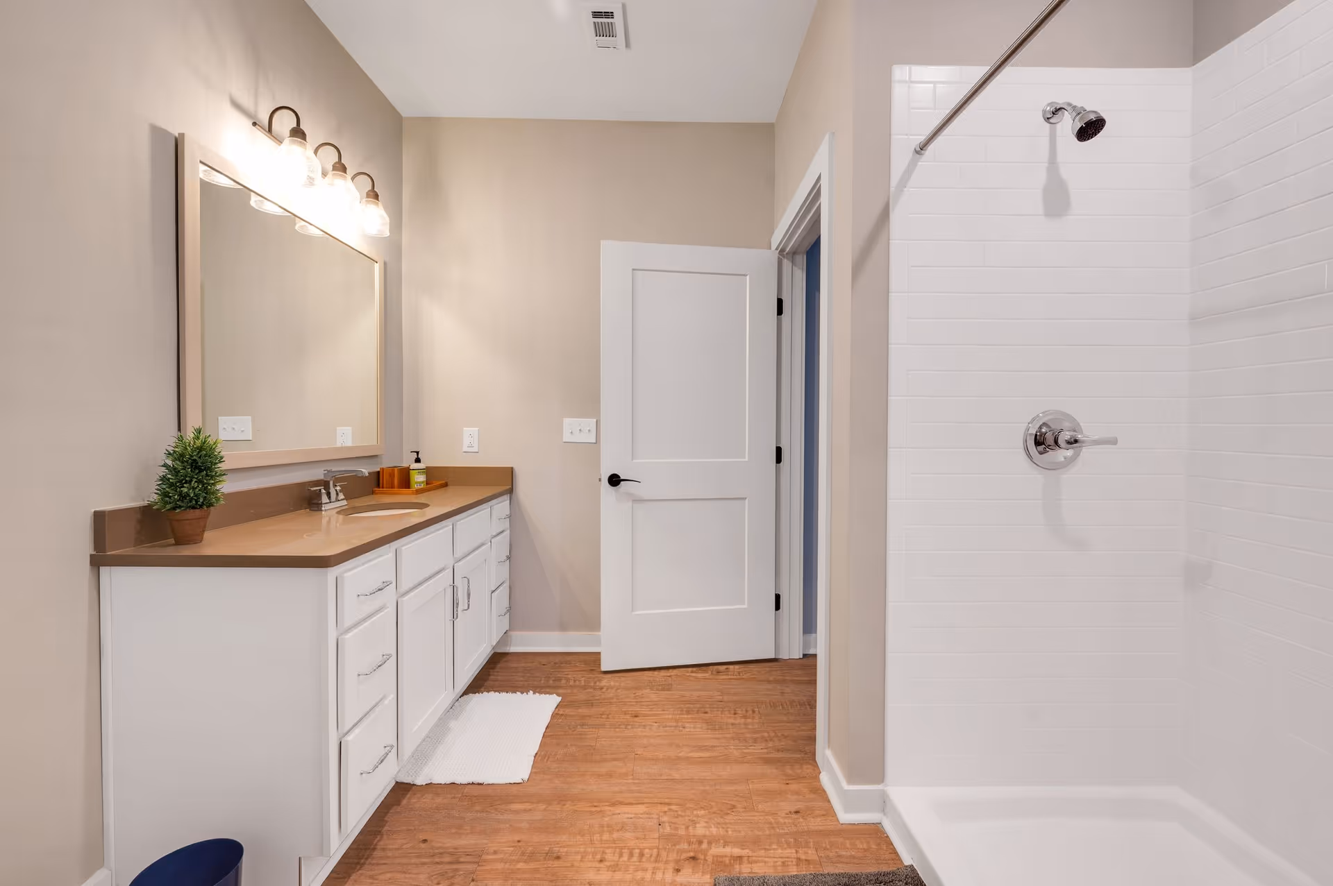 A clean and modern bathroom featuring a large vanity with a brown countertop, white cabinets, a rectangular mirror with three light fixtures above it, a small potted plant, soap dispenser, and a white door. To the right is a white tiled walk-in shower with a chrome showerhead and handle. The floor is wood-style, and there are two bath mats on the floor.