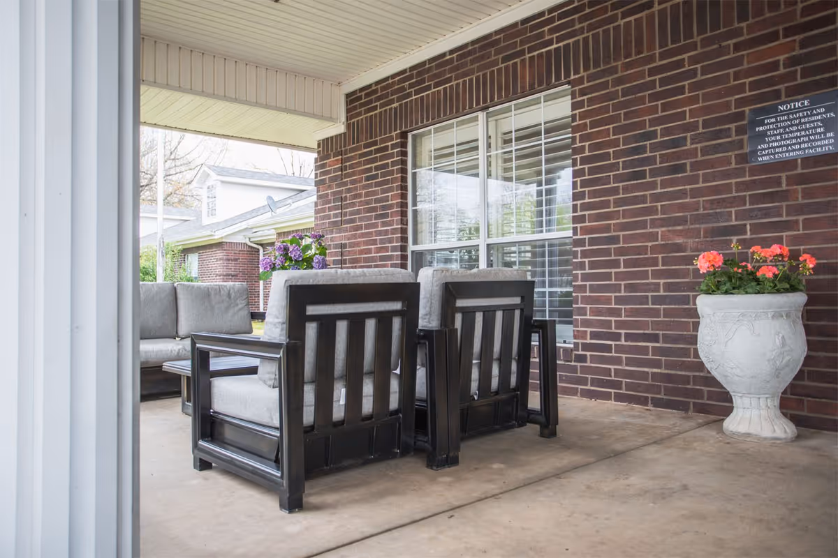 Covered outdoor patio area with cushioned seating including two armchairs and a sofa, a coffee table, a large window with white blinds on a brick wall, a white planter with pink flowers, and a notice sign on the wall.