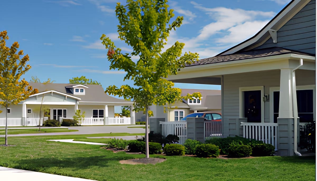 View of a senior living community with single-story houses featuring porches, surrounded by green lawns, small trees, and shrubs under a blue sky with some clouds.