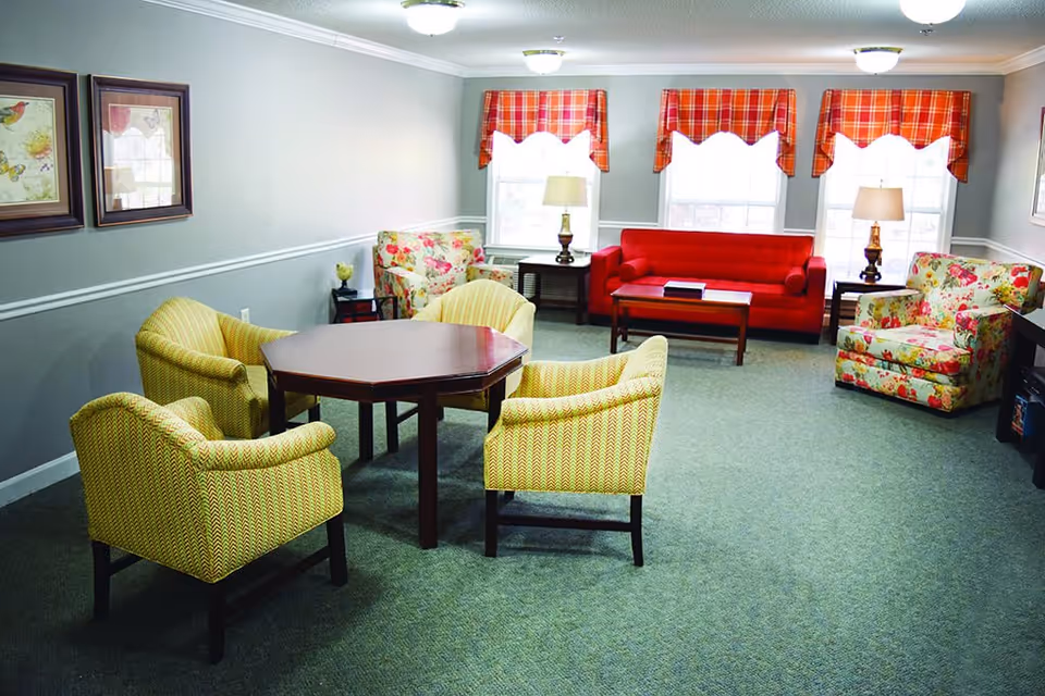 A cozy living room with a red sofa, two floral armchairs, and four yellow patterned chairs around a wooden octagonal table. The room has three windows with red plaid valances, two table lamps on dark wooden side tables, and framed butterfly artwork on the wall.